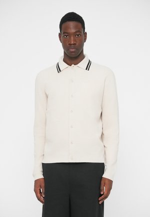 Polo štýlový pletený sveter v svetlej béžovej farbe s textúrovaným vzorom, s kontrastným čierno-bielym pásikavým golierom a predným zapínaním na gombíky.