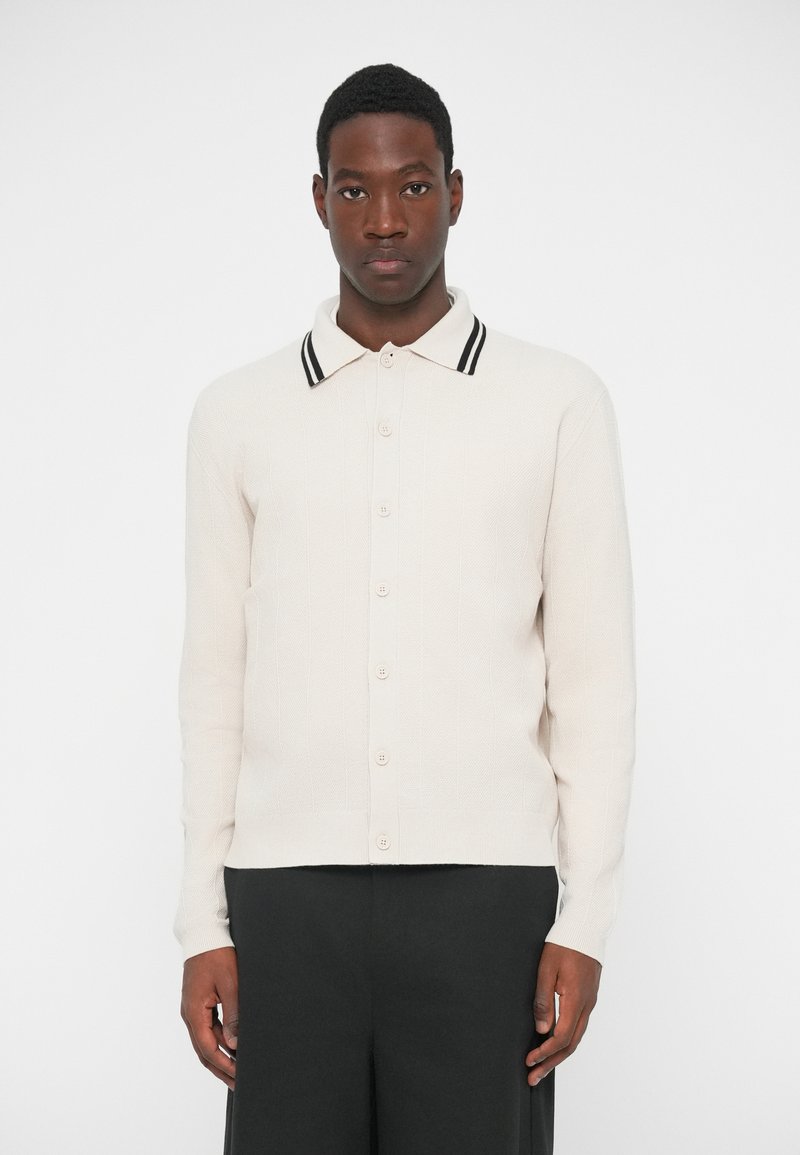 Polo stila adīts džemperis gaiši bēšā krāsā ar teksturētu rakstu, ar kontrastējošu melnbaltu svītrainu apkakli un pogu priekšpusi.