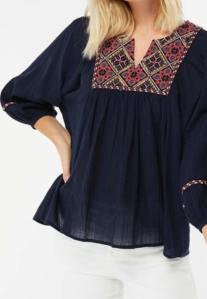 Marinebluse med farverig geometrisk broderi på bærestykke og manchetter, løse trekvartærmer, båret med hvide bukser.