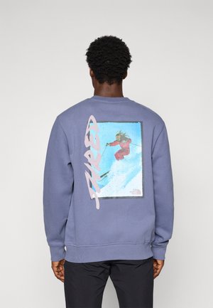 Man in een paarse sweatshirt met een groot beeld van een skiër die op de sneeuw springt en de tekst "BURTON" op de achterkant.
