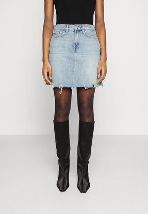 Femme portant une minijupe en denim bleu clair effilochée, des collants noirs transparents à pois, des bottes noires montantes et un haut noir sans manches, debout.