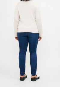 Maglione beige a trama con collo a giro, abbinato a jeans skinny blu scuro e scarpe con plateau nere. La vista da dietro mostra il taglio e il contrasto di colore.