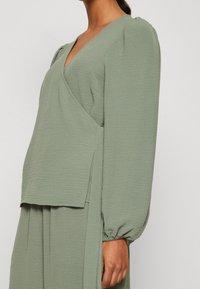 Abito verde oliva con scollo a V profondo e lunghe maniche a sbuffo. Il tessuto testurizzato presenta un design a strati con spacco laterale all'orlo.