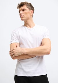 Νεαρός άνδρας με απλή λευκή μπλούζα, με σταυρωμένα τα χέρια, κοιτάζει προς τα αριστερά σε ουδέτερο φωτεινό φόντο.