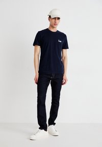 Marineblå bomulls-T-skjorte med en liten hvit logo, kombinert med mørke denimjeans og hvite sneakers, fullført med en lysfarget caps.