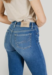 Femeie purtând blugi albaștri cu detalii din pietre strălucitoare pe buzunarele din spate și un top bej, tatuaje vizibile pe brațe, văzută de la mijlocul spatelui până la picioare.