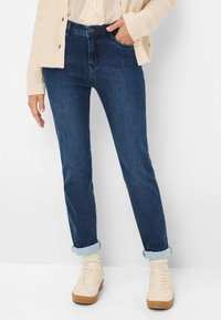 Jeans in jeans scuro con un design aderente, caratterizzati da risvolti sulle caviglie, passanti per la cintura e tasche frontali. Abbinati a sneaker alte beige.