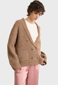 Bruine, chunky gebreide cardigan met dubbelknoopsluiting, diepe V-hals en grote voorklauwen, gedragen over een wit shirt en roze broek.