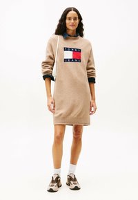 Frau in einem beigen Tommy Jeans Sweaterkleid, mit weißer Schultertasche, weißen Socken und beigen Turnschuhen, vor einem schlichten weißen Hintergrund stehend.