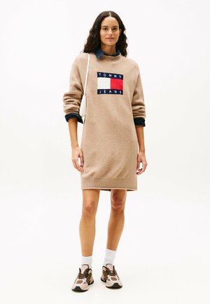 Vrouw in een beige Tommy Jeans sweaterjurk, met een witte schoudertas, witte sokken en beige sneakers, staand tegen een effen witte achtergrond.