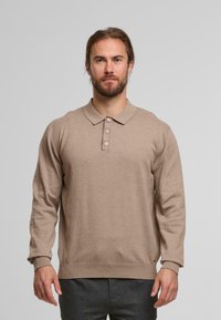 Man met een baard in een beige polo-shirt met lange mouwen en donkergrijze broek, staande tegen een effen lichtgrijze achtergrond.