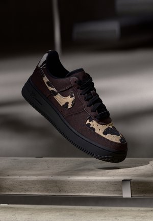 Schwarzer und brauner Turnschuh mit Kuhmuster-Akzenten auf dem Nike-Swoosh und der Spitze, schwebend über einer Betonoberfläche dargestellt.