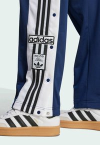 Close-up of navy and white Adidas track pants with black stripes and snap buttons worn with white Adidas Samba sneakers with gum soles.