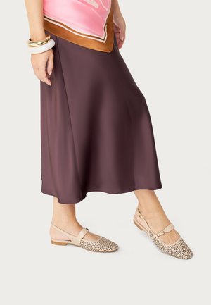 Mujer que lleva zapatos planos beige con talón abierto perforados, una falda midi marrón, una bufanda rosa y marrón, y múltiples pulseras en la muñeca izquierda.