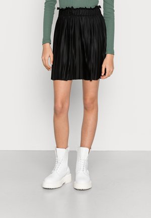 Personne portant une mini-jupe plissée noire, un haut vert à manches longues et des bottines blanches à lacets, debout sur un sol gris.