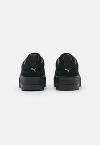 Schwarze Wildledersneaker mit einer dicken schwarzen Sohle. Besondere Merkmale sind ein goldenes Logo-Detail an der Ferse und ein strukturiertes Stoffdetail. Verstärkter Fersenbereich.