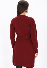 Hosszú, sötét hajú nő, aki övvel ellátott, hosszú ujjú bordó pulóverruhával és nagyméretű arany fülbevalókkal van öltözve, háttal áll.