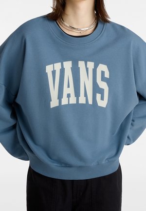 Person iført blå crewneck-sweatshirt med hvid "VANS" tekst og lagdelte sølvsmykker, kombineret med sorte bukser, på hvid baggrund.