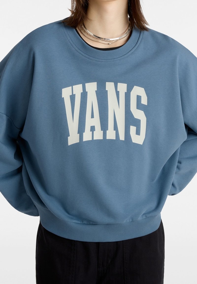 Personne portant un sweat-shirt bleu à col ras du cou avec le texte blanc "VANS" et des colliers argentés superposés, assorti à un pantalon noir, sur fond blanc.