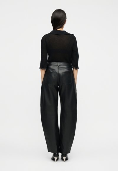 Γυναίκα που φοράει μαύρο διάφανο τοπ με ξεφτισμένα μανίκια, φορεμένο μέσα σε πλατύτερο δερμάτινο παντελόνι μαύρου χρώματος με ψηλή μέση, και μαύρες γόβες, με την πλάτη προς τα εμάς.