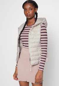 Helle beige, puffy Weste mit Kapuze, die einen Reißverschluss und Seitentaschen hat. Getragen über einem gestreiften Langarmshirt und einem geribbelten rosa Rock.