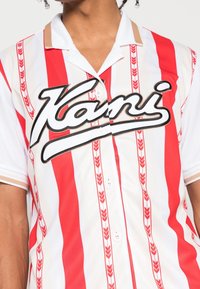 Camisa de manga corta a rayas rojas y blancas, hecha de un tejido ligero, con un logotipo negro bordado y patrones verticales decorativos.