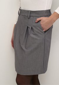 Personne portant une jupe grise longueur genou avec des plis sur le devant et des poches latérales, assortie à des collants noirs et un haut blanc à manches longues.