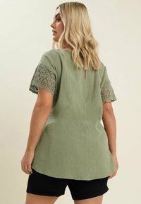 Olivgrön tunika med texturerat tyg, korta spetsärmar, nyckelhål bak och asymmetrisk fåll. Kombinerad med svarta shorts.