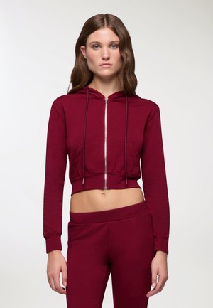 Crop hoodie bordeaux con zip frontale, cappuccio regolabile e tasca frontale; abbinato a pantaloni a vita alta coordinati, realizzati in tessuto morbido.
