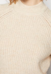 Pull beige en maille avec un col haut côtelé et des manches raglan. La texture présente un tissage serré avec des variations subtiles de teinte.