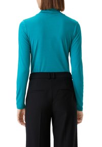 Turkusowa bluzka z długim rękawem i stójką, zestawiona z czarnymi, klasycznymi spodniami z przodu i z tyłu kieszeniami. Gładka tekstura.