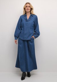 Completo in denim caratterizzato da un top a maniche lunghe con scollo a V arricciato e una gonna maxi coordinata, entrambi in una tonalità di blu medio uniforme.