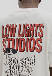 Cream-colored t-shirt featuring bold red and black text: "LOW LIGHT STUDIOS L.L.S." and "Represent yourself visually!" in varied fonts.