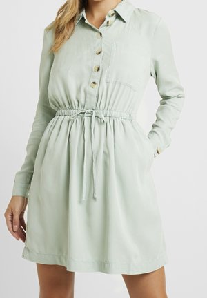 Lichtgroene jurk met lange mouwen, knopen aan de voorkant, een borstzak en een taille met trekkoord, gedragen door een persoon met één hand in de zak.