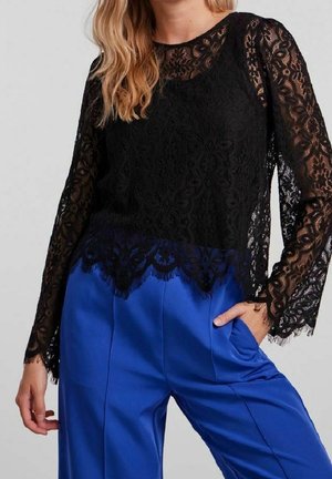 Vrouw die een zwarte kanten blouse met lange mouwen draagt, gecombineerd met felblauwe satijnen broek, hand in de zak, tegen een effen achtergrond.