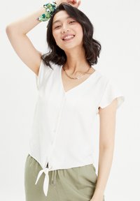 Blouse blanche à manches courtes avec un col en V et une fermeture par bouton, dotée d'un détail en nœud à l'ourlet et d'un tissu léger et texturé.