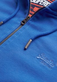 Sudadera azul con cremallera fabricada en suave felpa. Presenta herrajes plateados, logotipo bordado y diseño de forro interior en contraste. Costuras detalladas visibles.