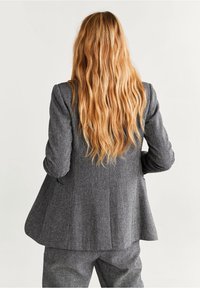 Blazer gris à motif avec une coupe ajustée, légèrement évasée à l'arrière, et une fente unique au dos. Le matériau semble texturé avec une finition lisse.