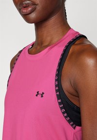 Różowy sportowy top bez rękawów z okrągłym dekoltem, z czarnymi ramiączkami ozdobionymi napisem "UNDER ARMOUR" oraz małym logo na piersi.