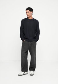 Pull en tricot côtelé bleu marine foncé, assorti à un jean noir ample et à des baskets blanches avec des rayures noires. La texture est marquée et décontractée.