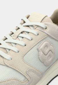 Közeli kép bézs és fehér sportcipőről, amelyen szarvasbőr és hálós panelek, fehér fűzők, valamint az oldalán és a nyelvén dombornyomott "BOSS" logó látható.