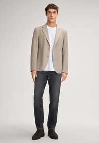 Blazer beige in misto lino con due bottoni, colletto a scatto e tasca applicata, indossato sopra una t-shirt bianca e jeans scuri con stivaletti marroni.
