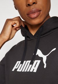 Čierna mikina s kapucňou a šnúrkami, na prednej strane je veľké biele logo Puma. Tkanina vyzerá hladko s matným povrchom. Detailný pohľad na výstrih a tvár.