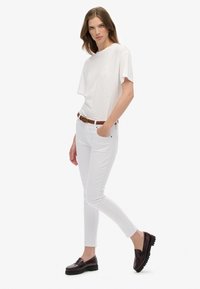 Camiseta blanca de manga corta, jeans ajustados blancos, cinturón trenzado marrón y mocasines burdeos oscuros con suela texturizada. Un conjunto simple y casual.