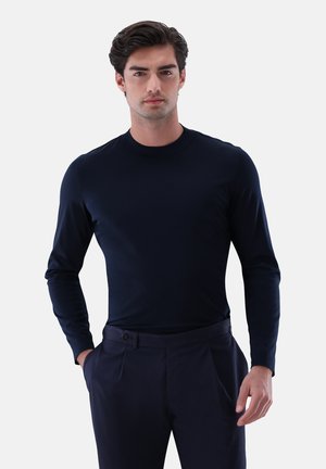 Dunkelblaues, langärmliges, eng anliegendes Shirt mit Rundhalsausschnitt. Der Stoff wirkt glatt und leicht und wird mit maßgeschneiderten blauen Hosen kombiniert.