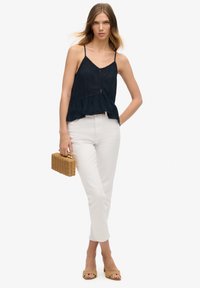 Marineblauwe peplumtop met dunne bandjes, witte aansluitende broek en beige blokhak sandalen. Model met een kleine geweven boodschappentas.