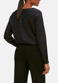 Blusa nera a maniche lunghe con una texture liscia, dettaglio del plissè sul retro e scollo rotondo, abbinata a pantaloni neri sartoriali.