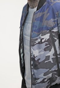 Kurtka bomber w kamuflażu w odcieniach niebieskiego, z zamkiem błyskawicznym, prążkowanym kołnierzykiem i kieszeniami bocznymi. Pod spodem szara koszulka z nadrukowanym tekstem.