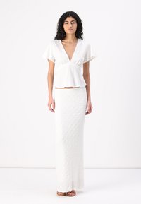 Hvit peplum-bluse med korte volangermer, kombinert med en lang hvit teksturert skjørt. Modellen står oppreist og har på seg flate sandaler.
