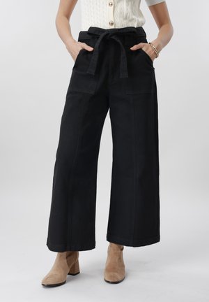 Ženska, ki nosi črne široke hlače z žepi spredaj in zavezanim pasom, kombinirane s konjak rjavimi gležnjaki in belim zgornjim delom s sponkami.